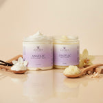 Two jars of 'Angelic' whipped body cream and body polish on a beige surface with vanilla beans and flowers.