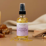 Bottle of Cocoa Cashmere body oil with a label on a wooden surface with candles and stones.