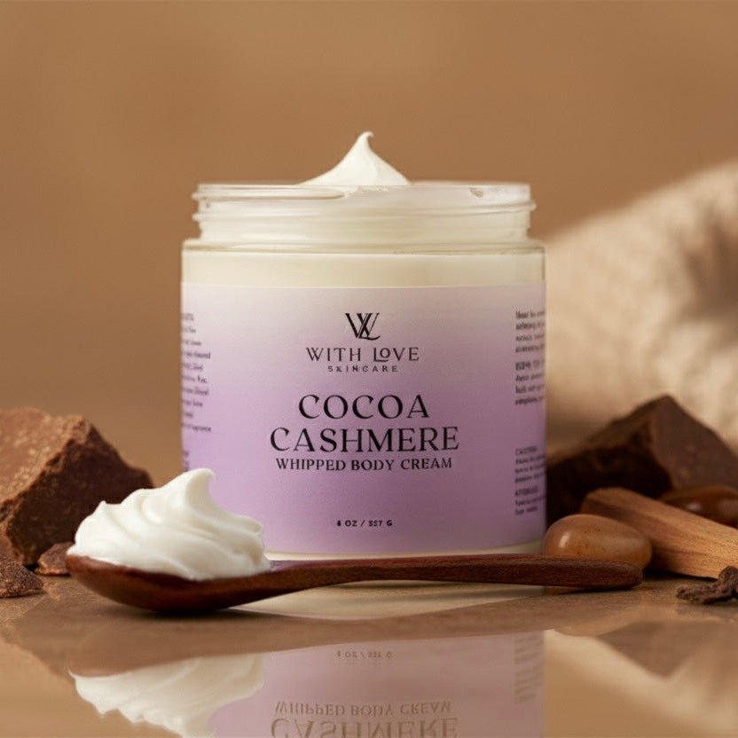 Jar of cocoa cashmere whipped body cream with chocolate pieces and a wooden spoon on a reflective surface.