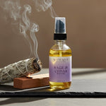 Bottle of Sage & Cedar body oil with a smoking sage bundle and candle on a dark surface.